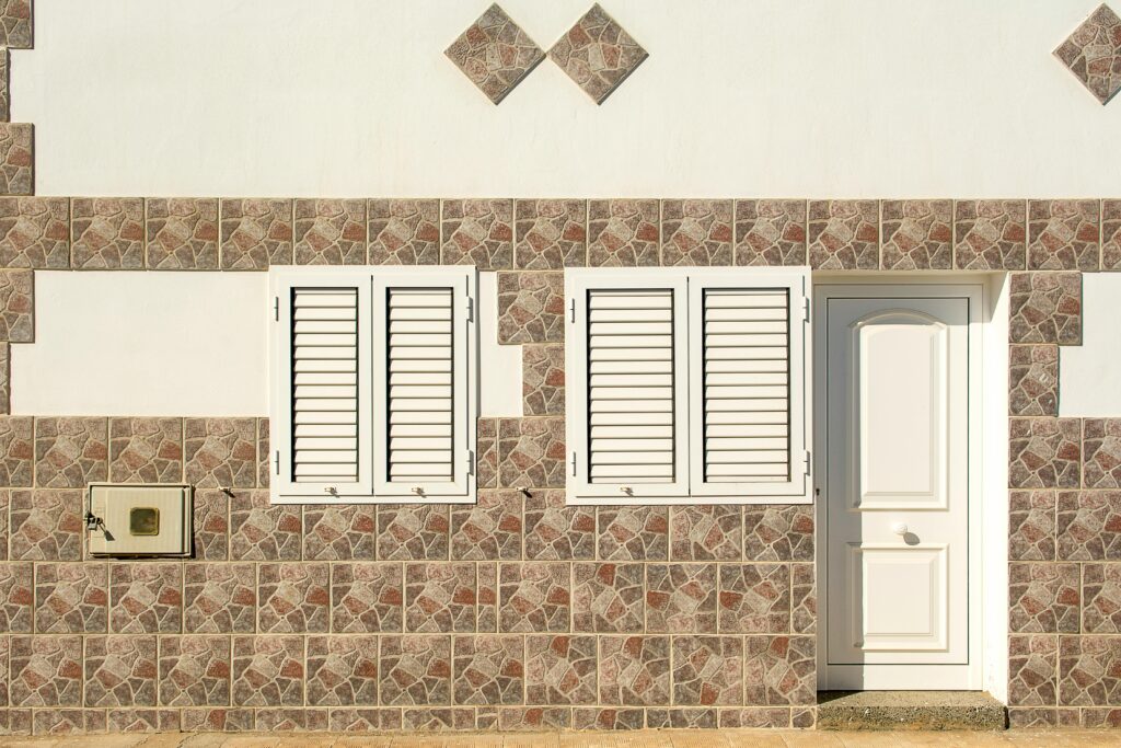 Close-up of a building facade featuring two white shuttered windows and a white door set within a mosaic tile wall.